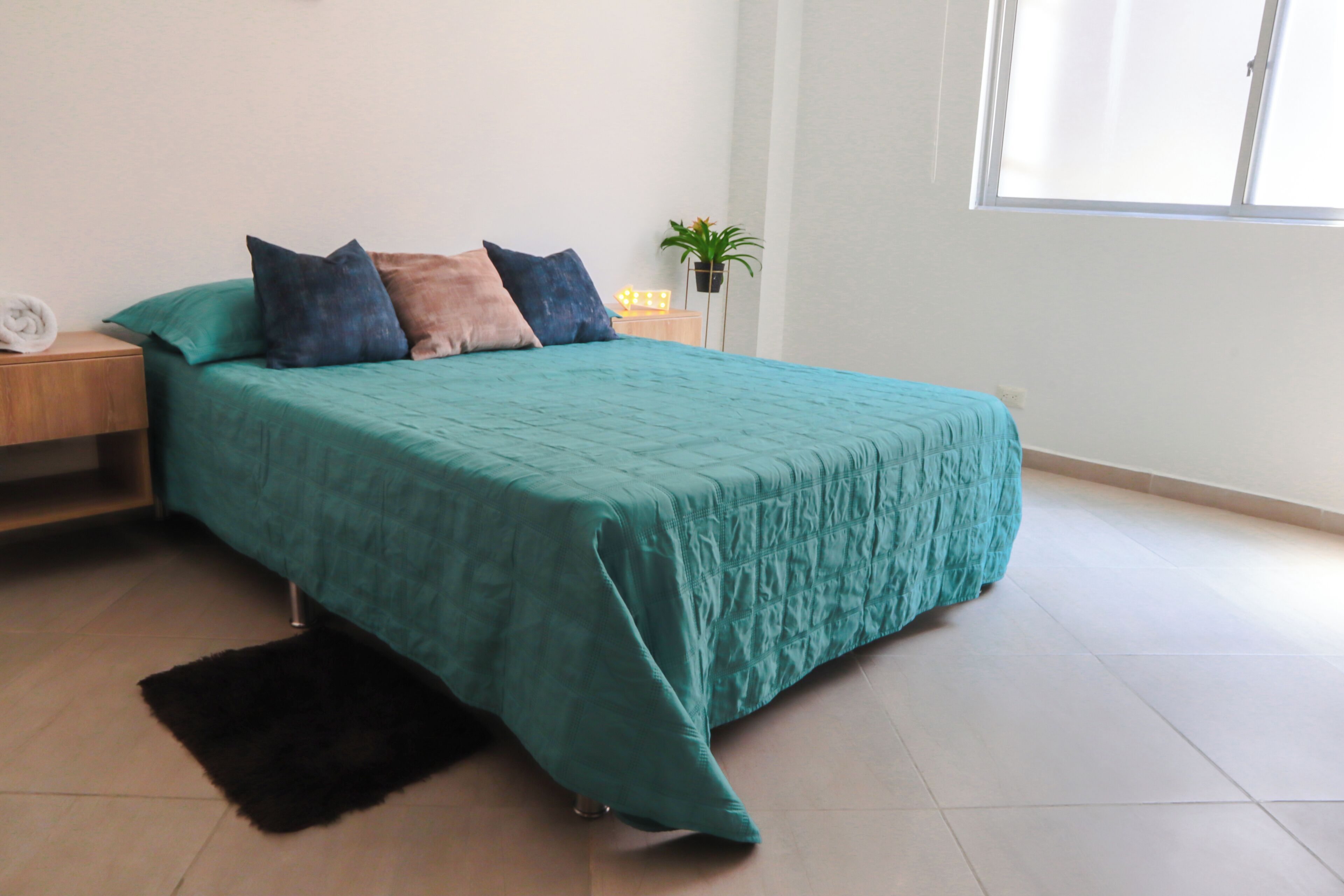 A neatly made bed with a teal bedspread and multiple pillows, a small bedside table with a lamp, and a potted plant on a nightstand.