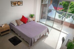 A bedroom with a bed, a nightstand, a small rug, a potted plant, and a large window. - Manila Apartaestudios  (Medellín)