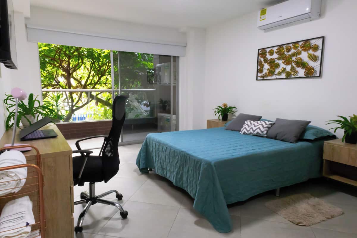 A bedroom with a bed, a desk with a chair, a window with a view of trees, and a wall-mounted air conditioner.