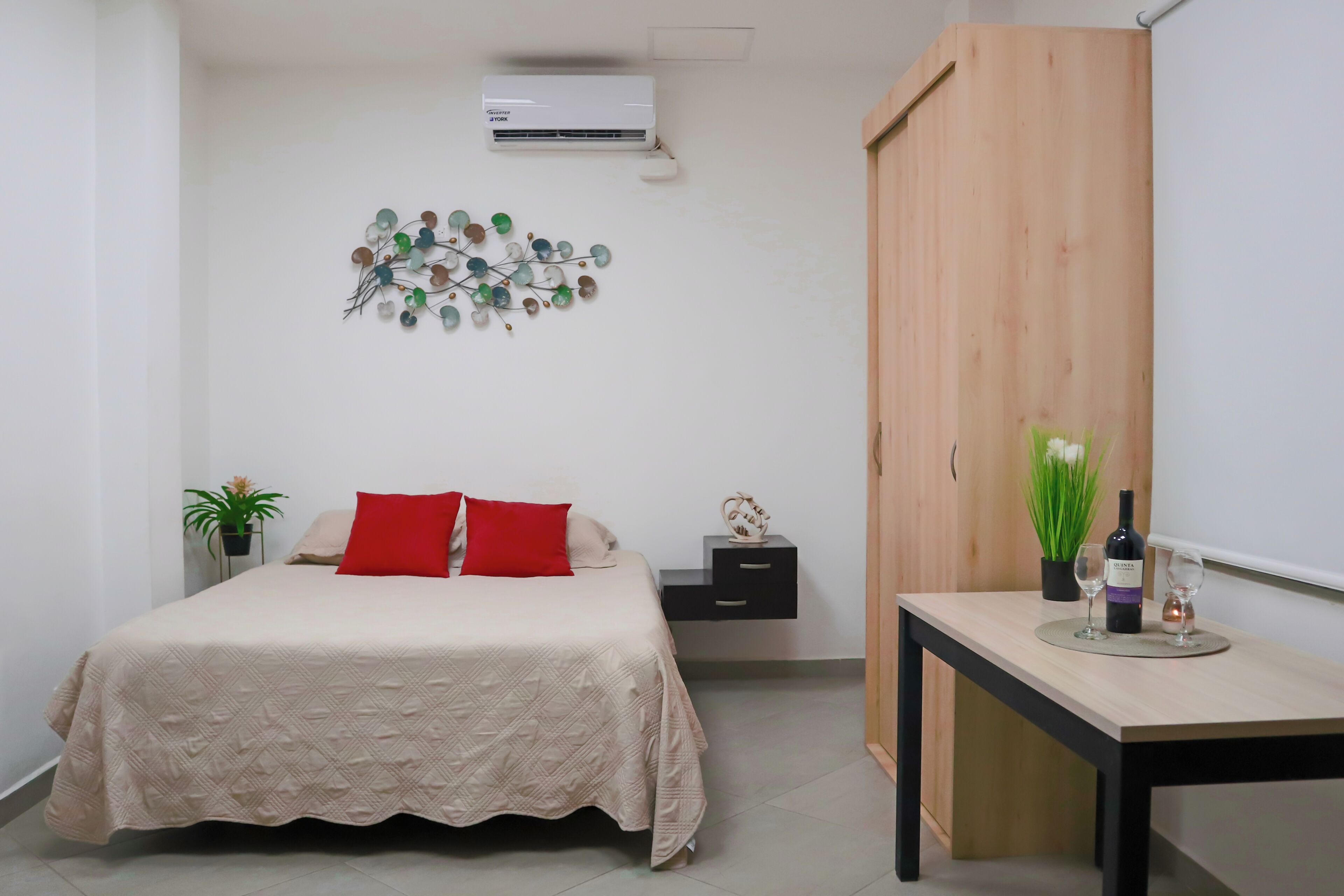 A bedroom with a bed, a nightstand, a desk, a wardrobe, a wall-mounted air conditioner, and a decorative wall piece.