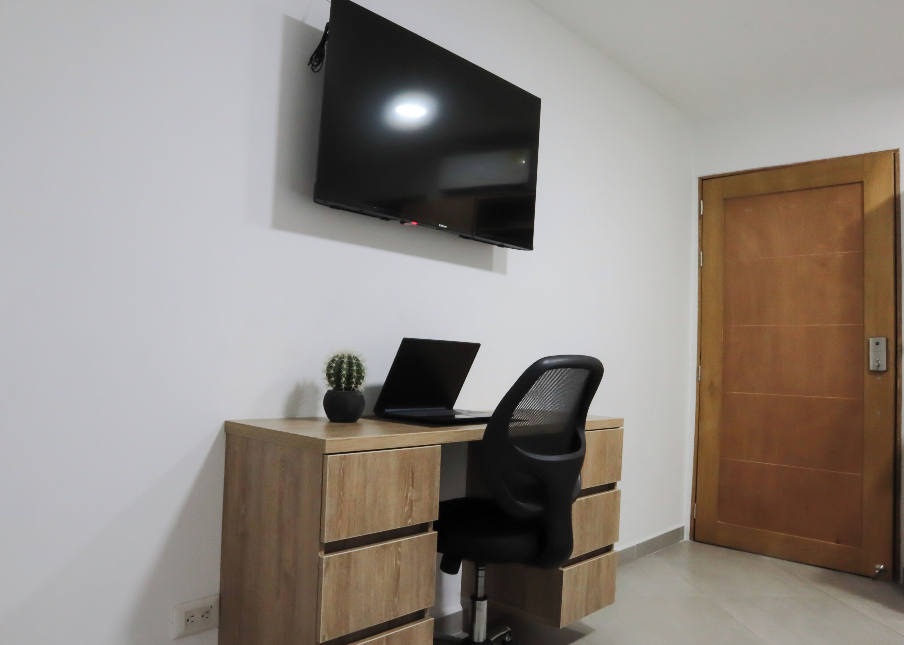 Une pièce avec un bureau en bois, une chaise noire, un téléviseur fixé au mur et une porte en bois.