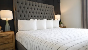 A neatly made bed with a tufted headboard, two bedside lamps, and a wooden nightstand with drawers. - Xhale Resort and Spa (Idaho Falls)