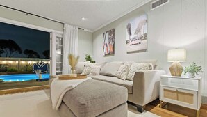 Una sala de estar moderna con un sofá, un reposapiés y una mesita de noche con una lámpara. Se ve una ventana grande con vista a una piscina y una cancha de baloncesto.