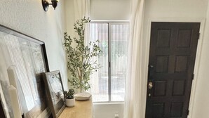 Una habitación con una mesa de madera, cuadros enmarcados, una planta en maceta, una ventana con cortinas y una puerta oscura.