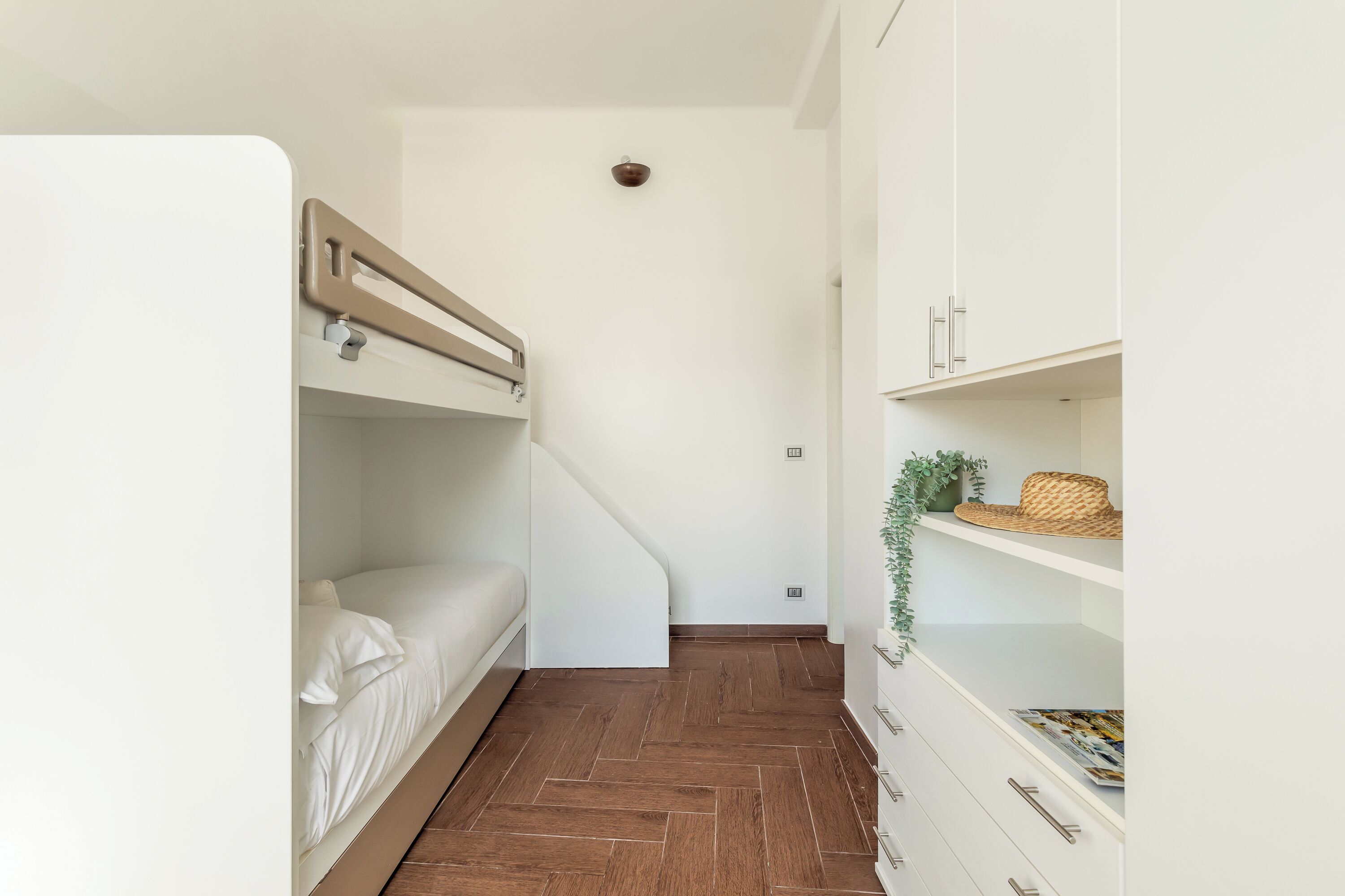 A compact room with a bunk bed, a white wardrobe with drawers, and a herringbone-patterned floor.