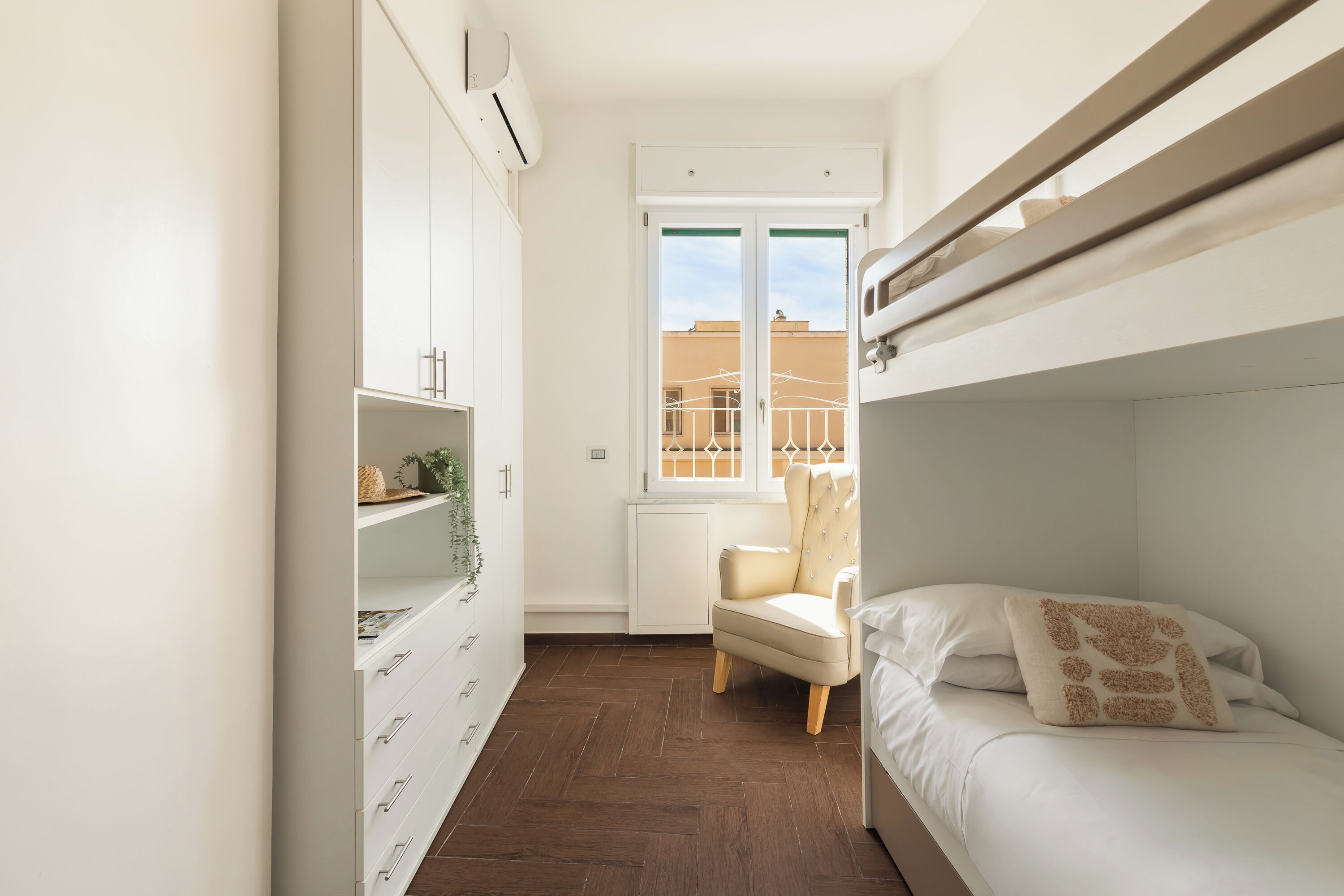 A compact bedroom with a bunk bed, a white wardrobe with drawers, a chair, and a window with a view of buildings.