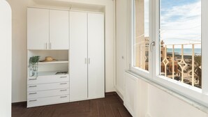 Une pièce avec une armoire blanche, une petite étagère sur laquelle se trouvent un chapeau et une plante, et une fenêtre donnant sur des immeubles et un ciel dégagé.