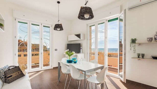 Un salon moderne avec une table à manger en verre, des chaises blanches et un canapé. On y trouve deux lampes suspendues noires, un téléviseur et une plante accrochée au mur.
