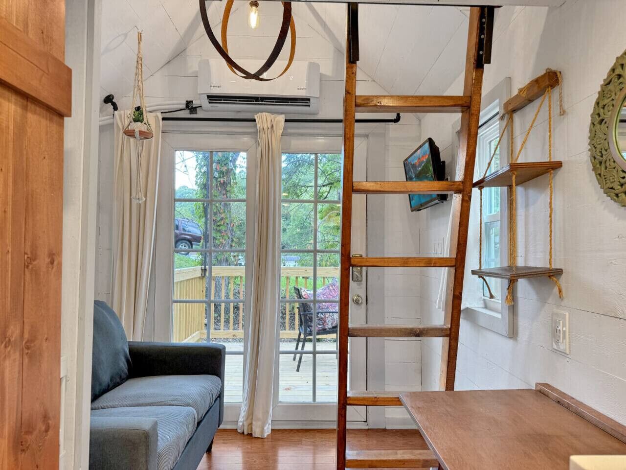 Una habitación con piso de madera, sofá gris, escalera, televisor en un estante y vista a una terraza y follaje exterior.