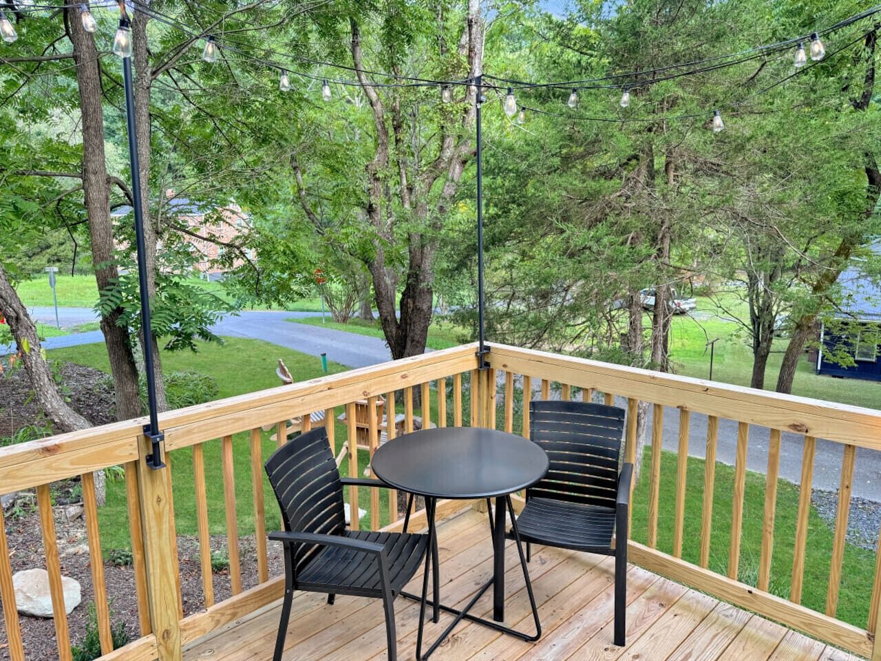 A wooden deck with a round table and two chairs, surrounded by trees and a view of a road.