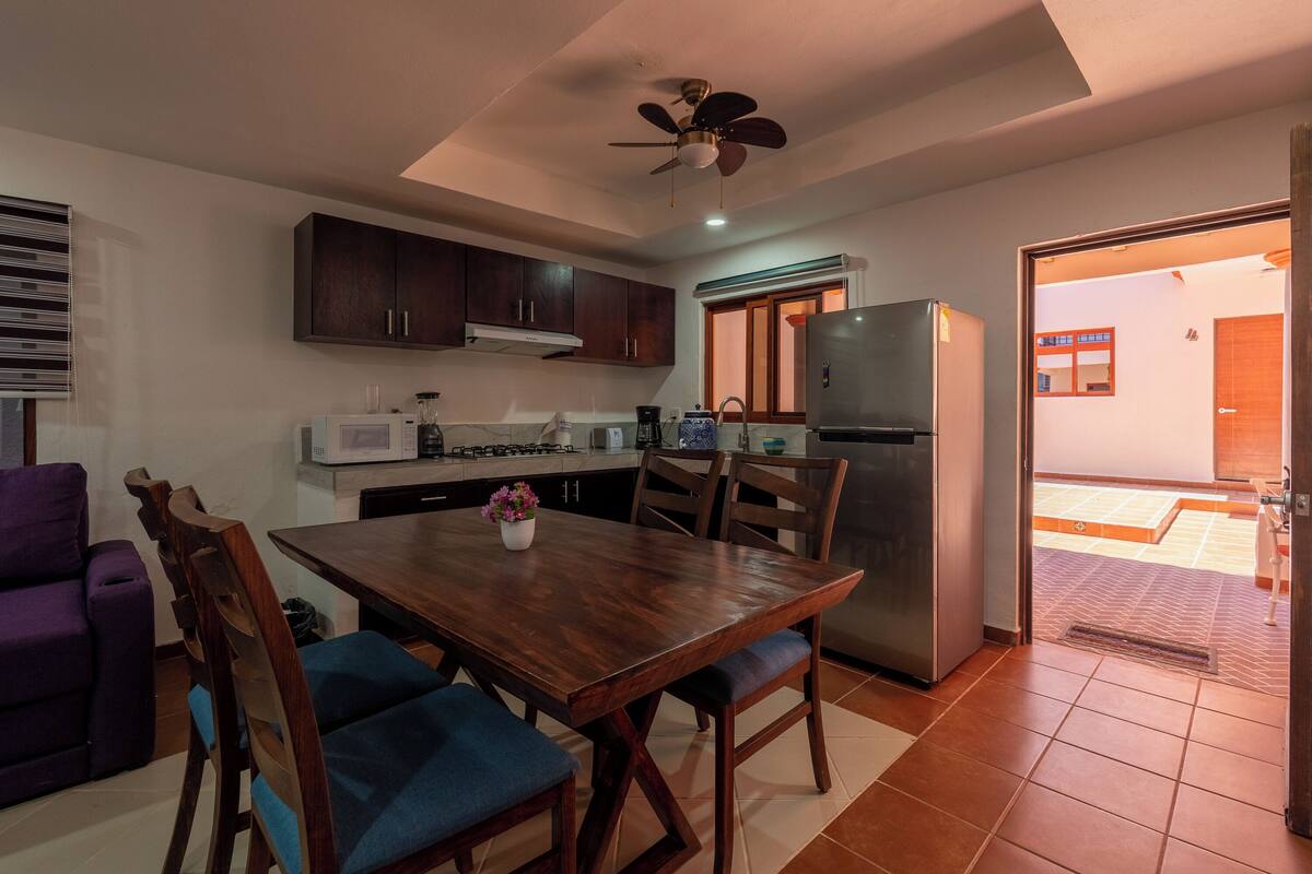 A modern kitchen with dark wooden cabinets, a stainless steel refrigerator, and a dining area with a wooden table and blue chairs.