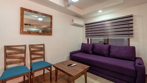 Un salon avec un canapé violet, deux fauteuils en bois, une petite table en bois, un ventilateur de plafond, un miroir et un appareil de climatisation.