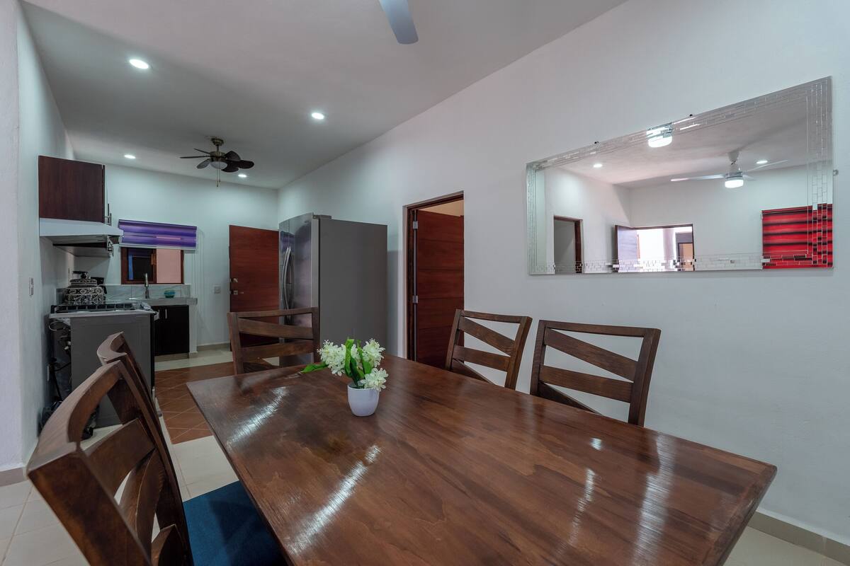 A dining area with a wooden table, chairs, and a potted plant. There is a kitchen in the background with cabinets and appliances.