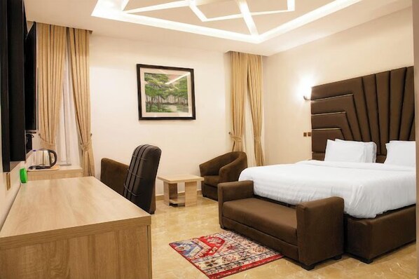 A hotel room with a bed, two armchairs, a desk, and a coffee table. There is a framed picture on the wall and a window with curtains.