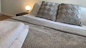 Ein ordentlich bezogenes Bett mit grauer Steudecke und Kissen, eine Nachttischlampe und Holzboden.