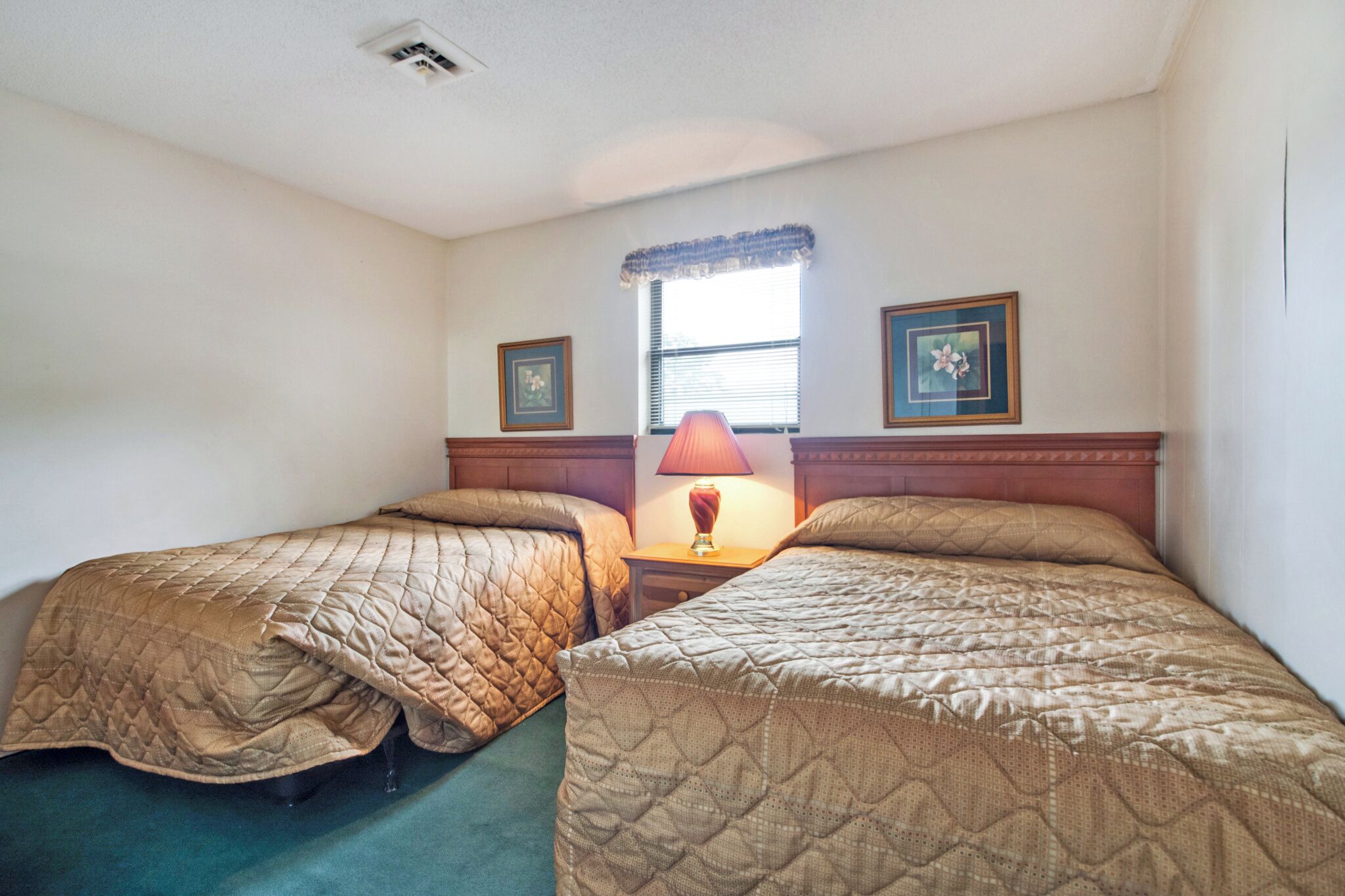 A room with two beds, a window with blinds, a lamp, and two framed pictures on the wall.