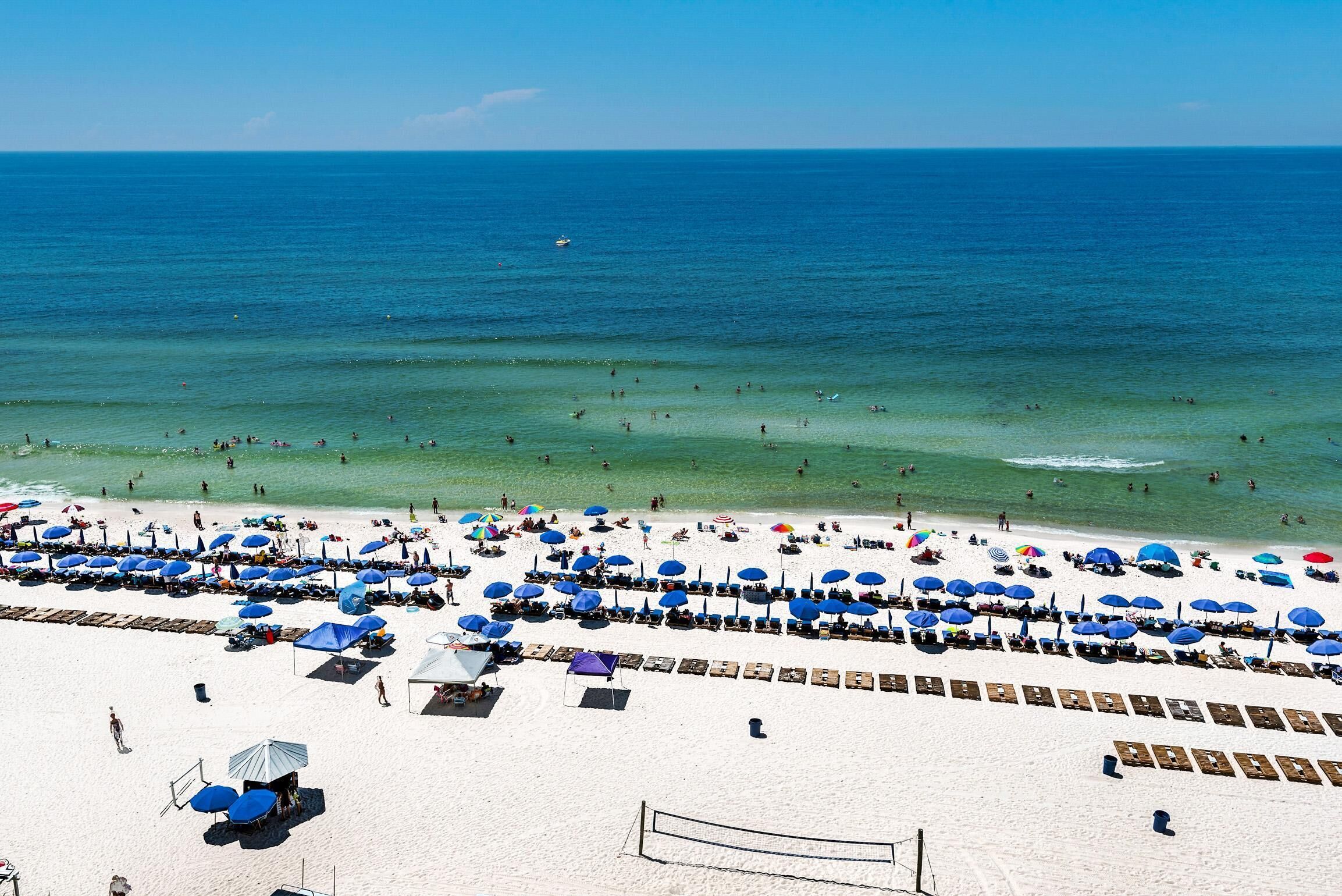 En strand med rækker af liggestole og parasoller, et volleyballnet og havet i baggrunden.