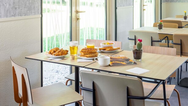 A breakfast table with croissants, waffles, and a drink, set in a room with large windows and a view of greenery. - Ibis Budget Bourg-En-Bresse (Bourg-en-Bresse)