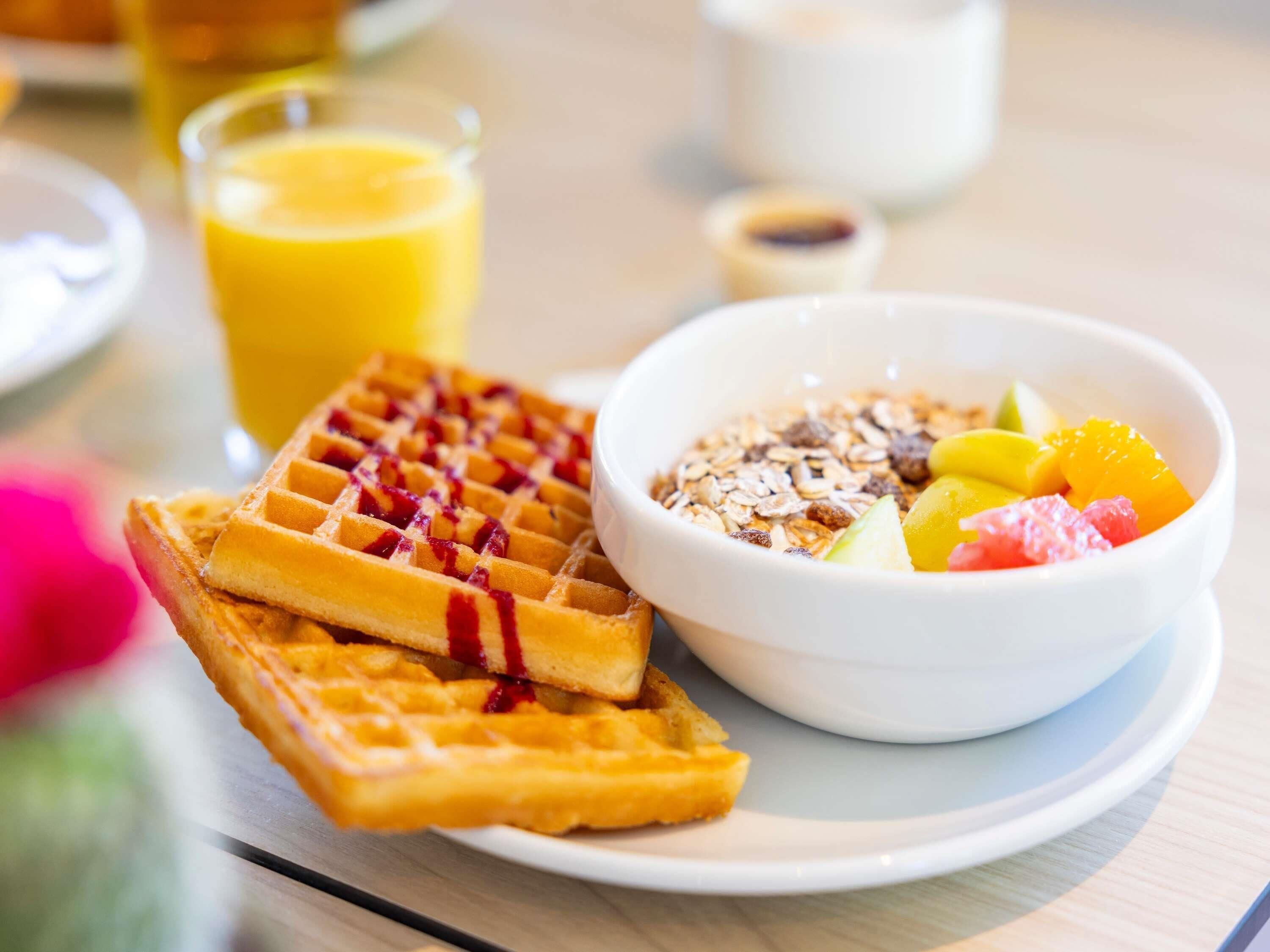 A breakfast setting with waffles, a bowl of fruit and granola, and a glass of orange juice.