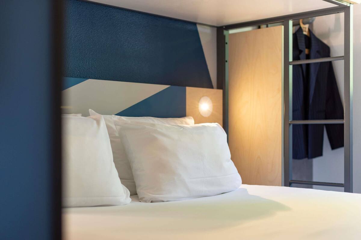 A modern hotel room with a bed, white pillows, a geometric wall design, and a wooden wardrobe with hanging clothes.