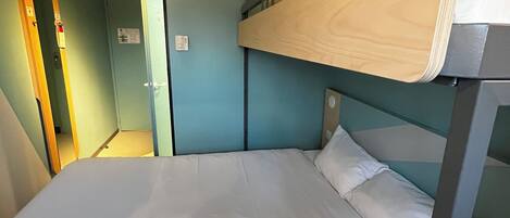 Una cama litera con cabecera de madera, un colchón blanco y una manta de color claro. Se observa una unidad de aire acondicionado fijada a la pared sobre la cama y una puerta a la izquierda.