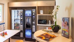 A modern hotel room with a minibar, a microwave, a coffee maker, a table with a magazine, and a small plant. - Ibis Budget Bourg-En-Bresse (Bourg-en-Bresse)