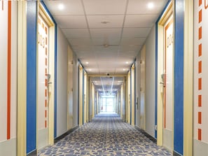 A hotel hallway with doors marked with room numbers, a patterned carpet, and a ceiling with recessed lighting. - Ibis Budget Bourg-En-Bresse (Bourg-en-Bresse)