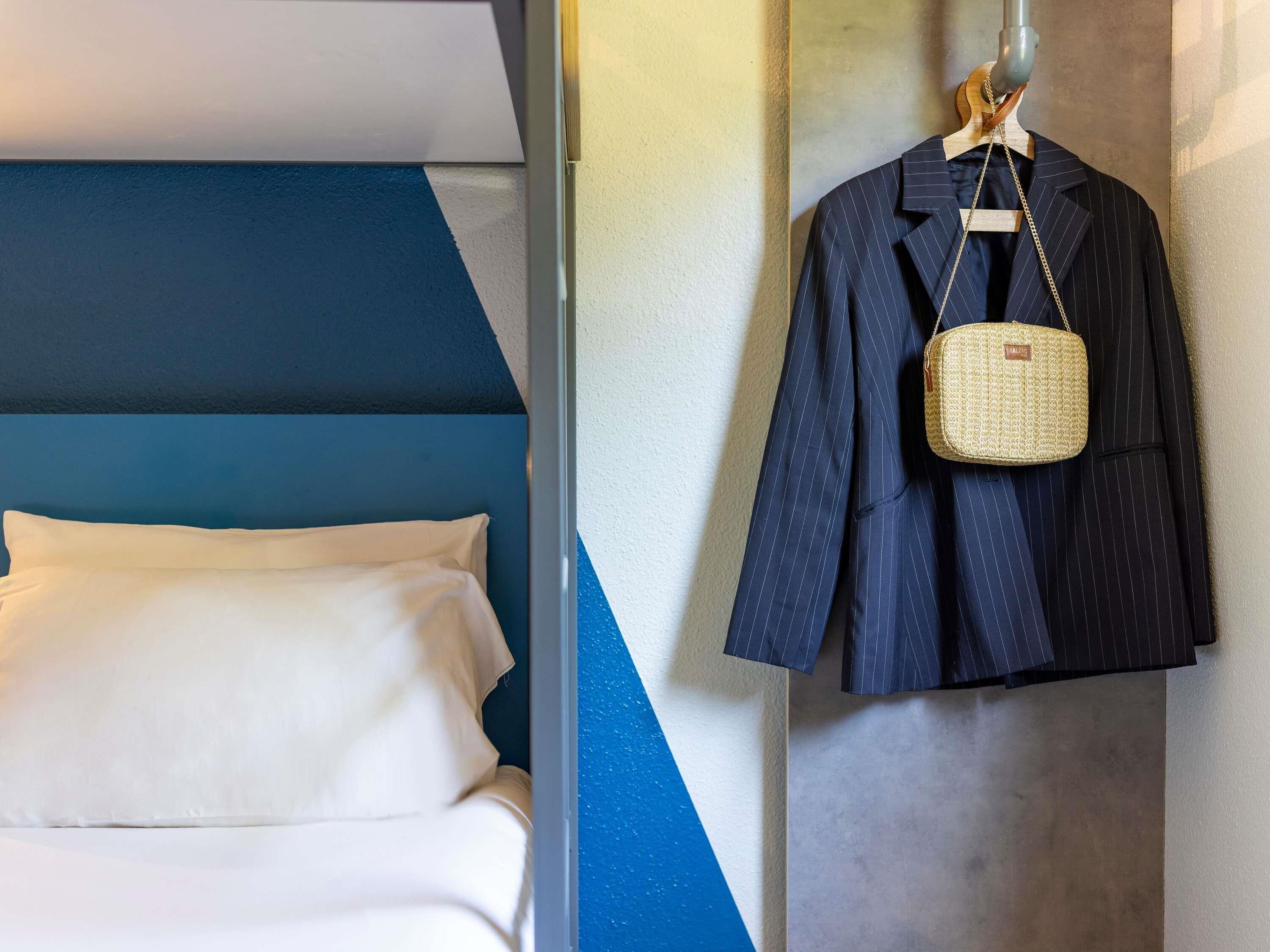 A hotel room with a bed, a hanging coat rack with a pinstriped suit and a woven bag, and a geometric wall design.