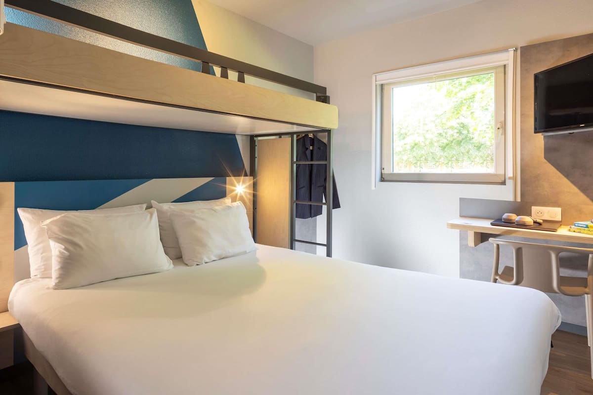 A hotel room with a bunk bed, a desk with a chair, a TV, and a window with a view of greenery.