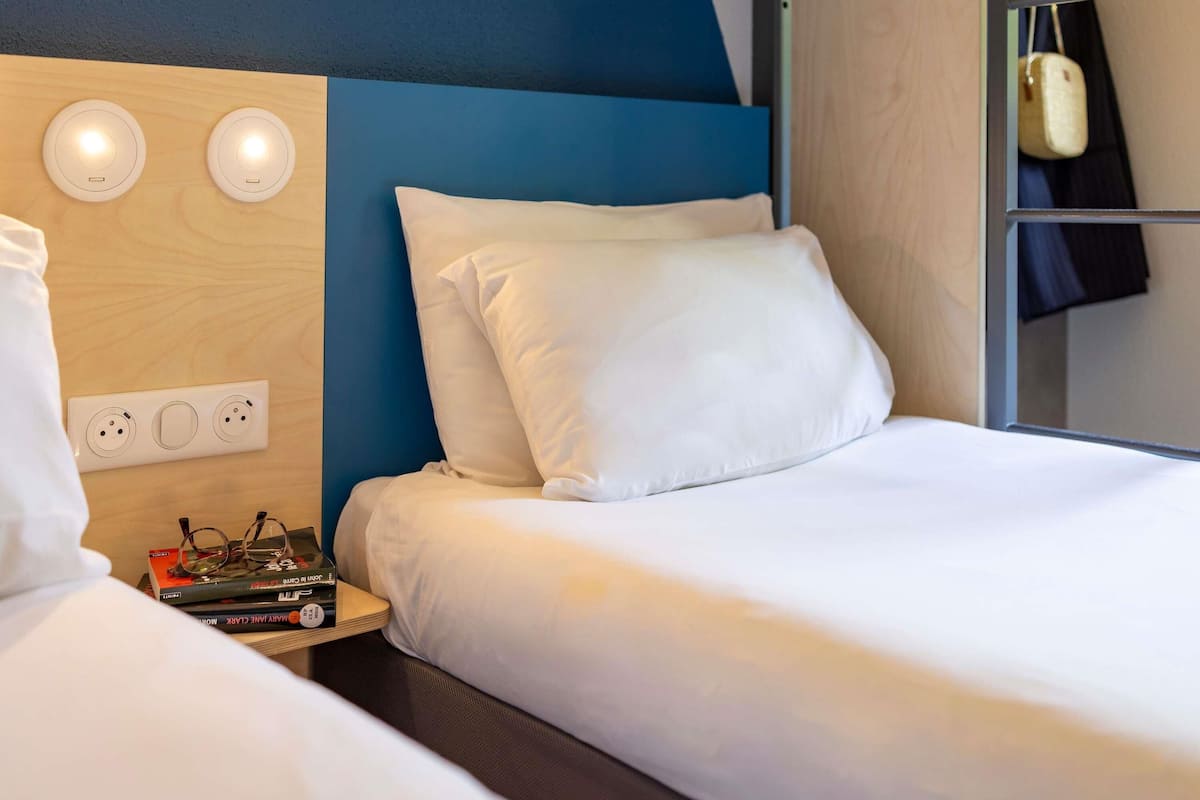 A hotel room with a bunk bed, a bedside table with glasses and a book, a power outlet, and a wall-mounted light fixture.