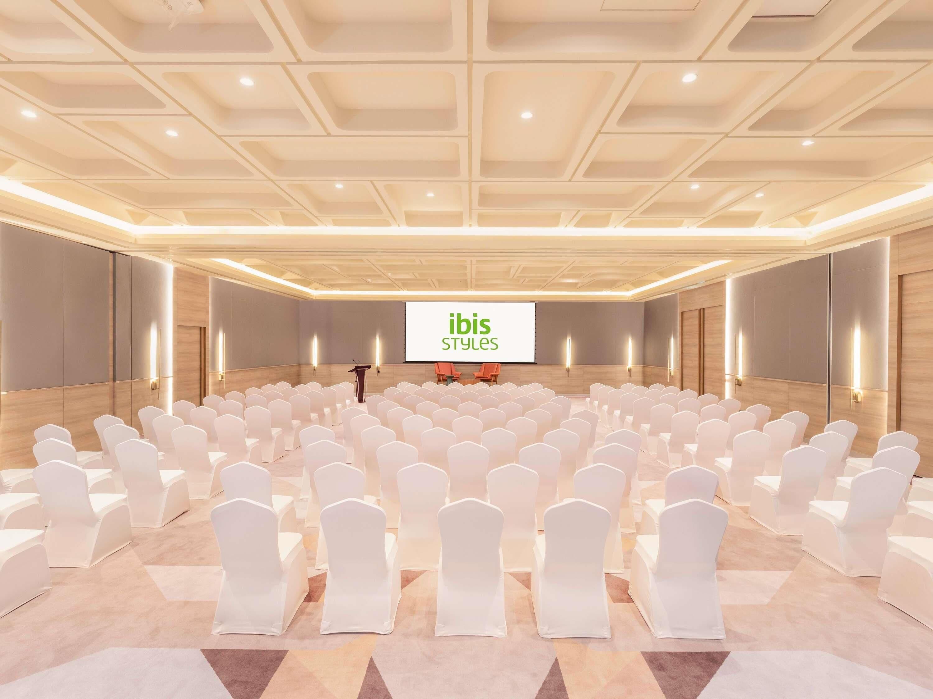 A conference room with rows of white chairs, a podium, and a screen displaying 'ibis Styles'.