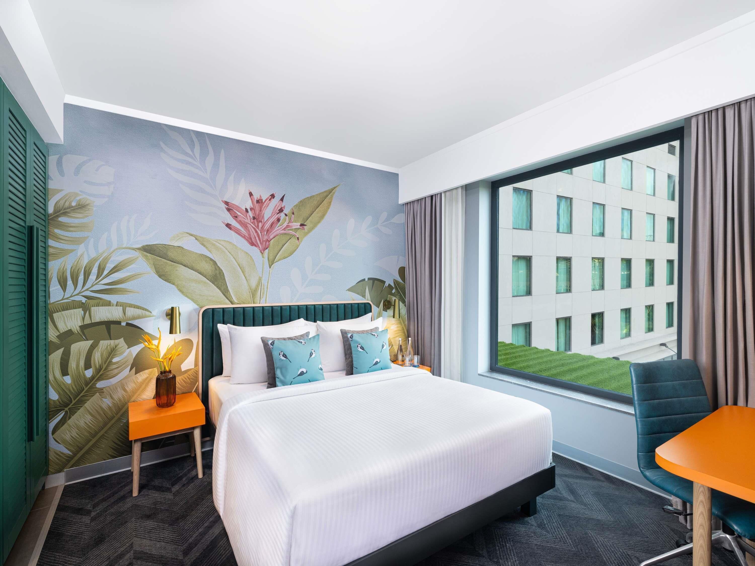 A hotel room with a bed, a desk, a chair, a window with a view of a building, and a wall with a tropical mural.