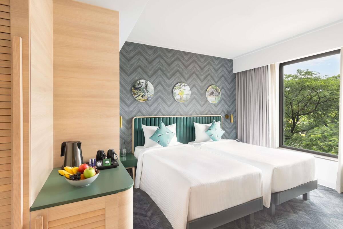 A hotel room with a bed, a small desk with a fruit bowl, and a view of trees through the window.
