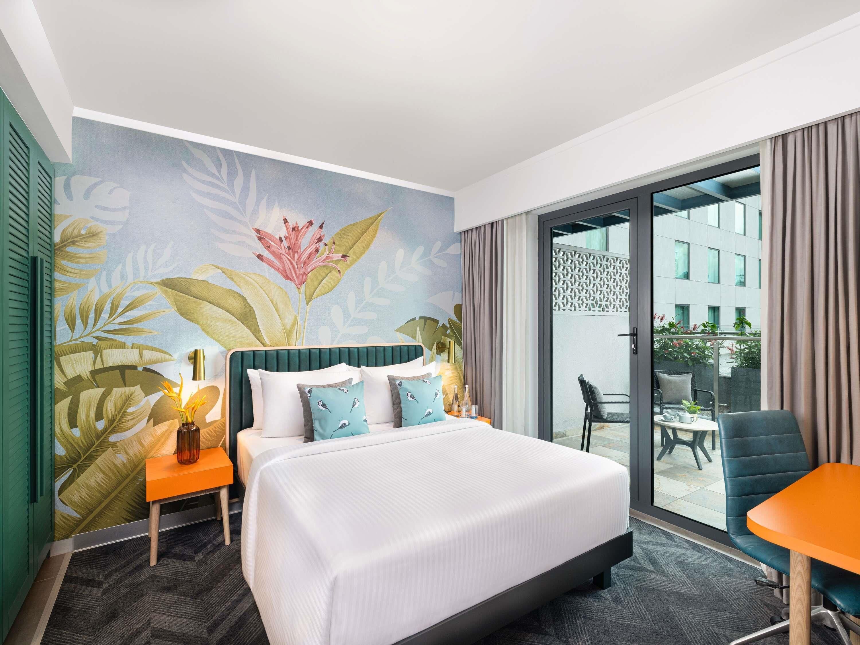 A hotel room with a large bed, a small table, a chair, and a balcony with a view of buildings.