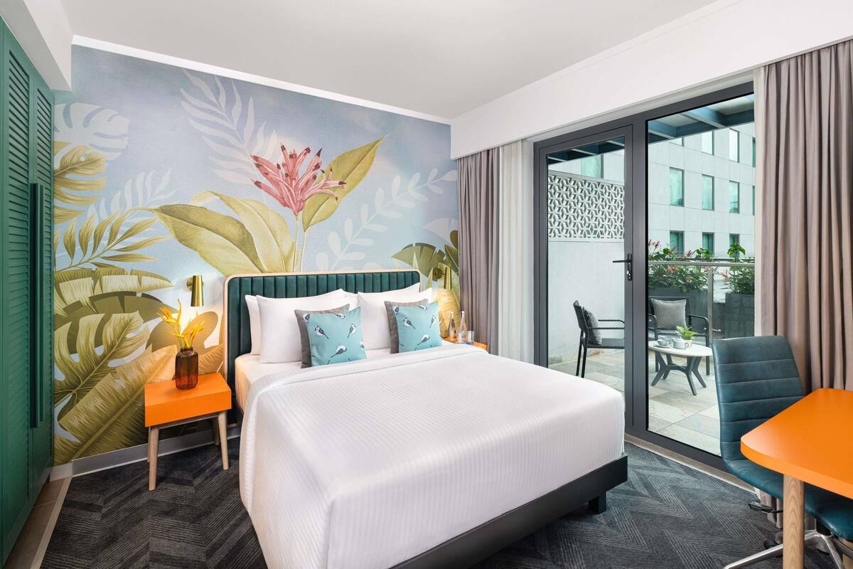 A hotel room with a large bed, a small table, a chair, and a balcony with a view of buildings.