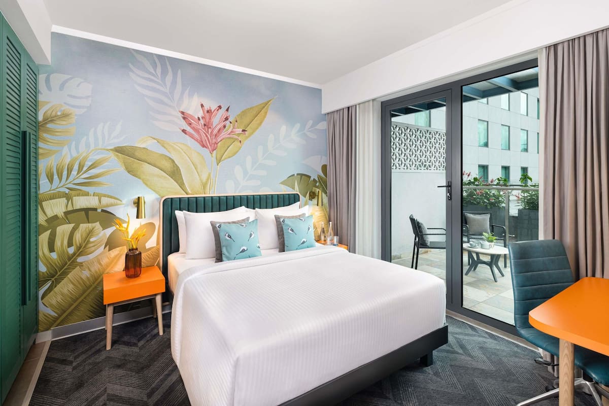 A hotel room with a large bed, a small table, a chair, and a balcony with a view of buildings.