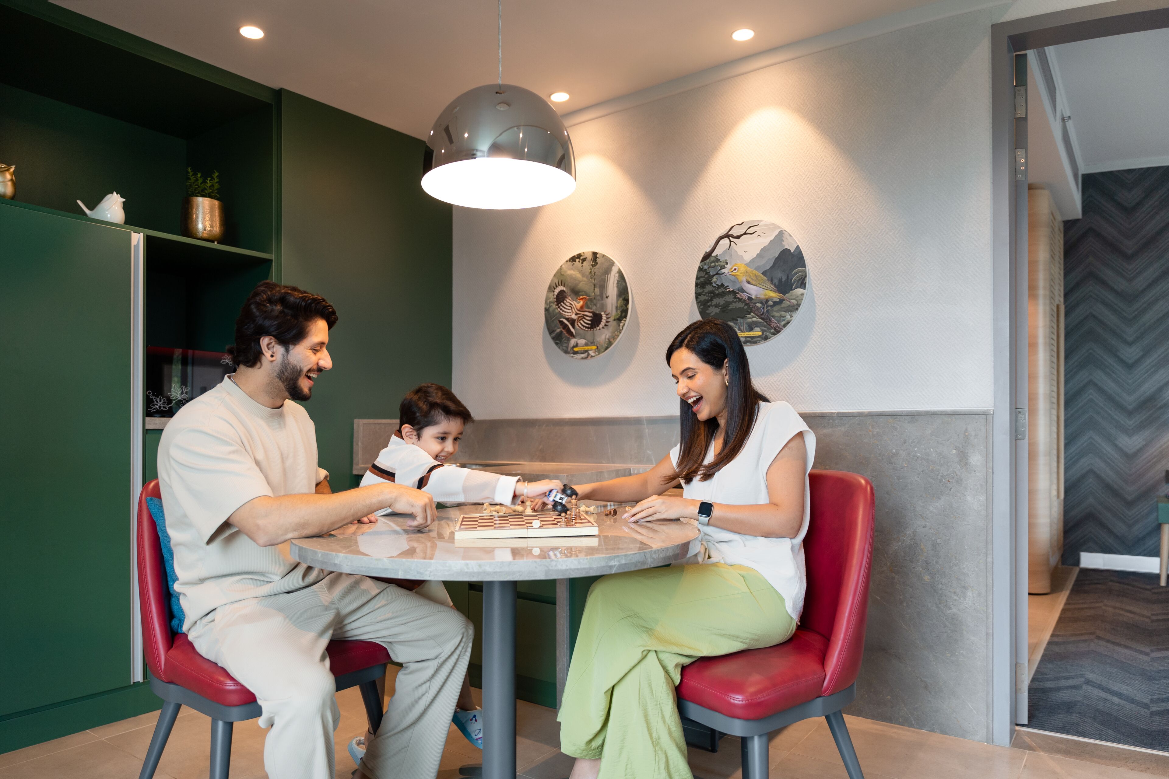 Una familia jugando ajedrez en una mesa redonda dentro de un ambiente moderno, con paredes verdes y detalles decorativos.