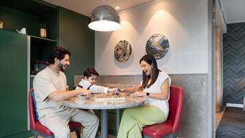 Una familia jugando ajedrez en una mesa redonda dentro de un ambiente moderno, con paredes verdes y detalles decorativos.