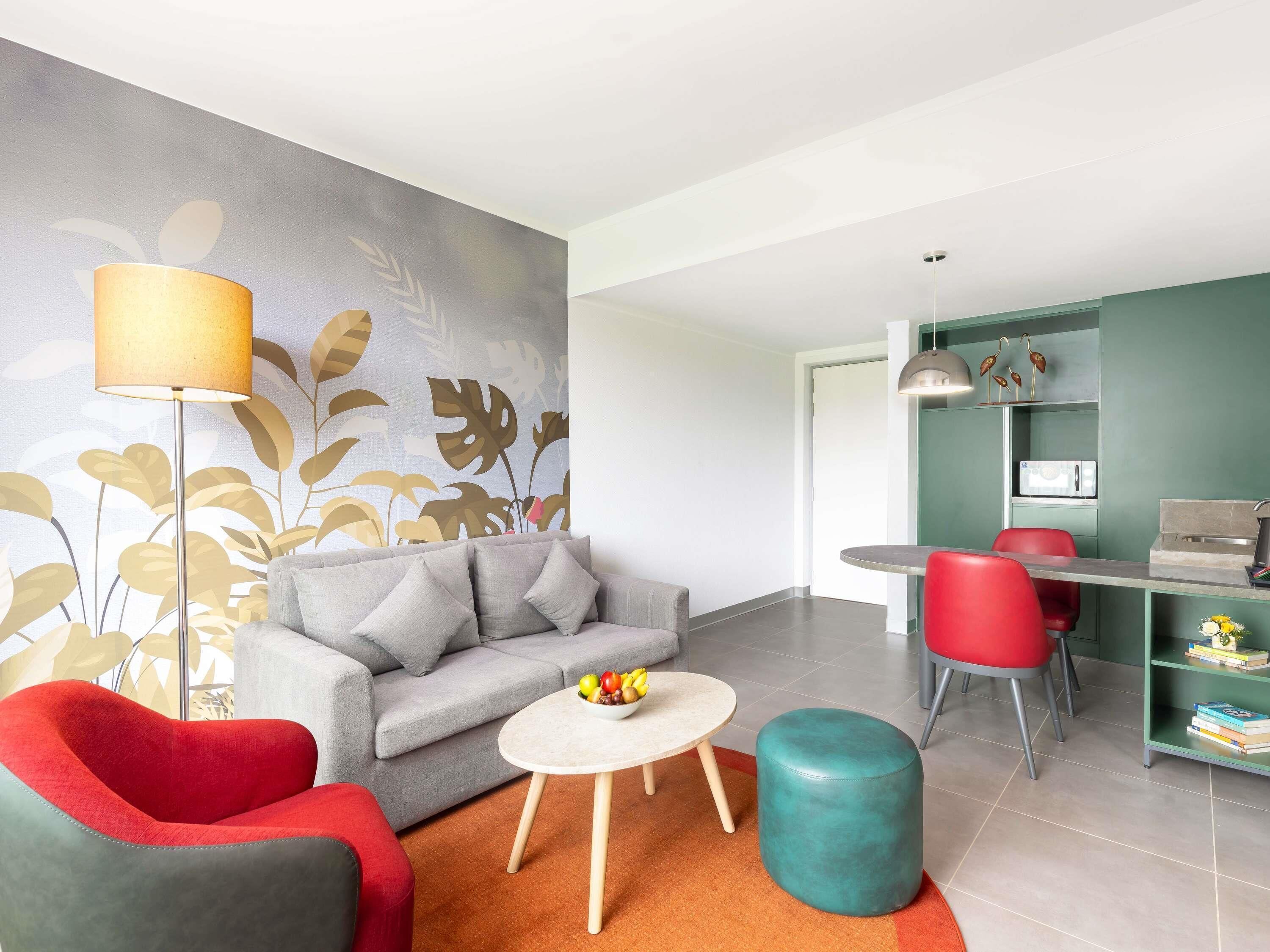 Una sala de estar moderna con un sofá gris, un sillón rojo, un reposapiés color turquesa y una mesita redonda con fruta.