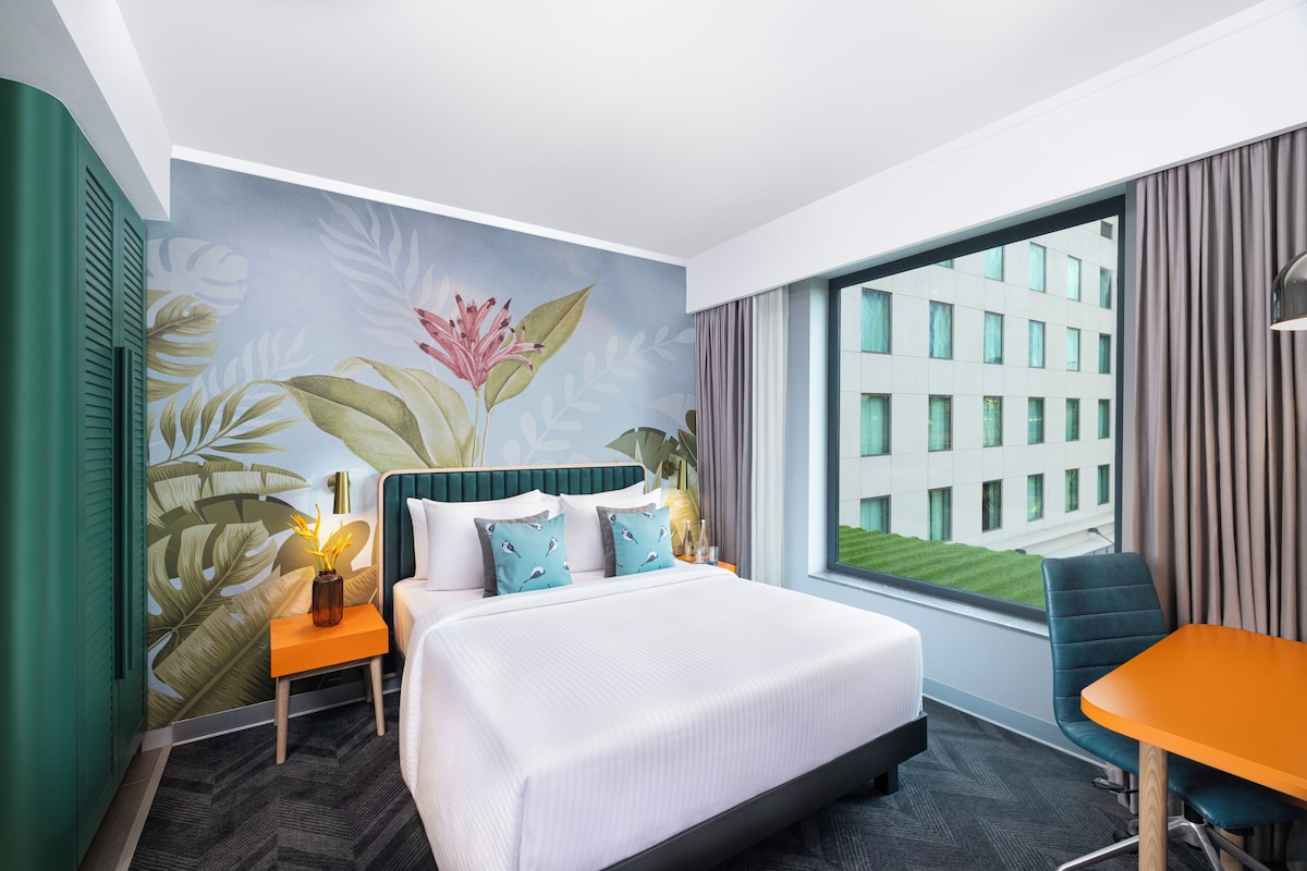 A hotel room with a bed, a small table, a chair, a nightstand, a lamp, and a window with a view of a building.