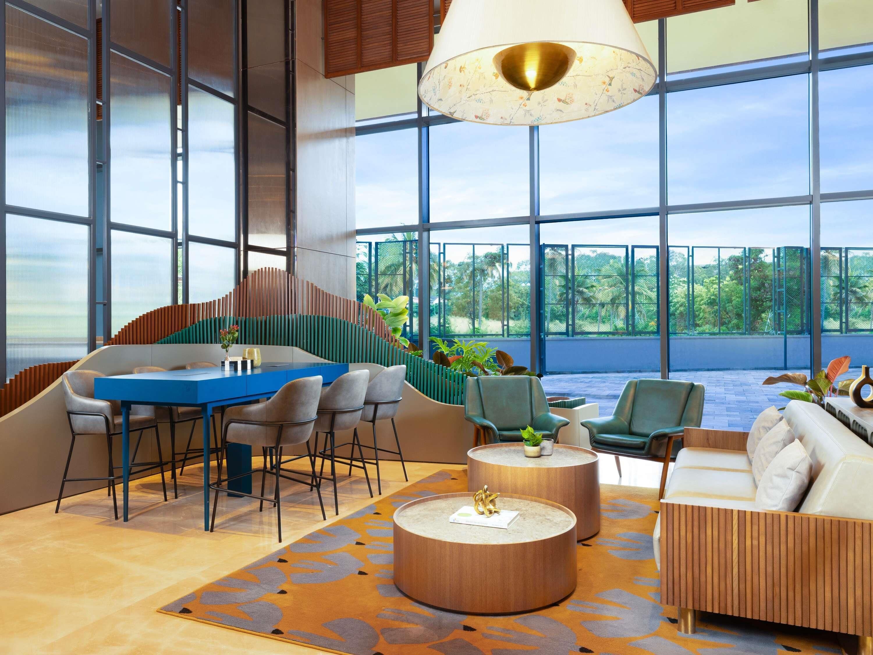 A modern interior with a blue dining table, multiple chairs, a round coffee table, and a sofa. Large windows offer a view of the outdoors.