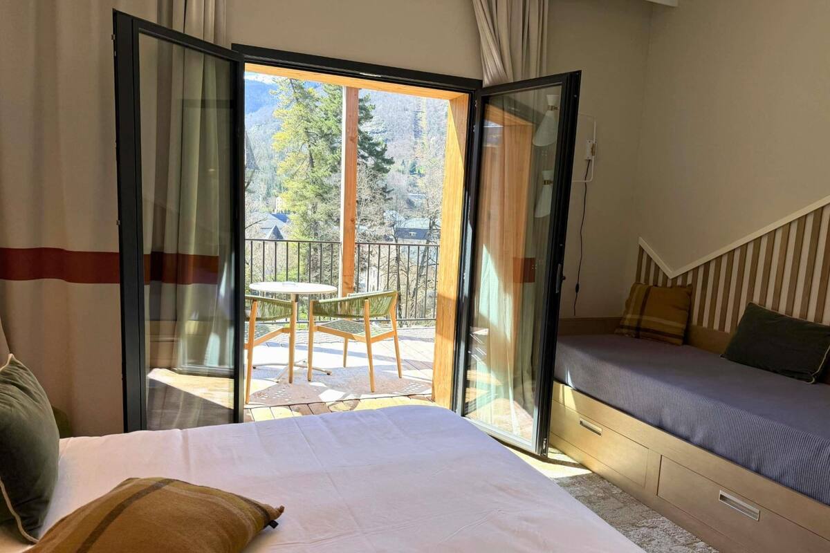 Ein Schlafzimmer mit Bett, einer Couch, einem Balkon mit Tisch und Stühlen und Blick auf Bäume und Häuser.