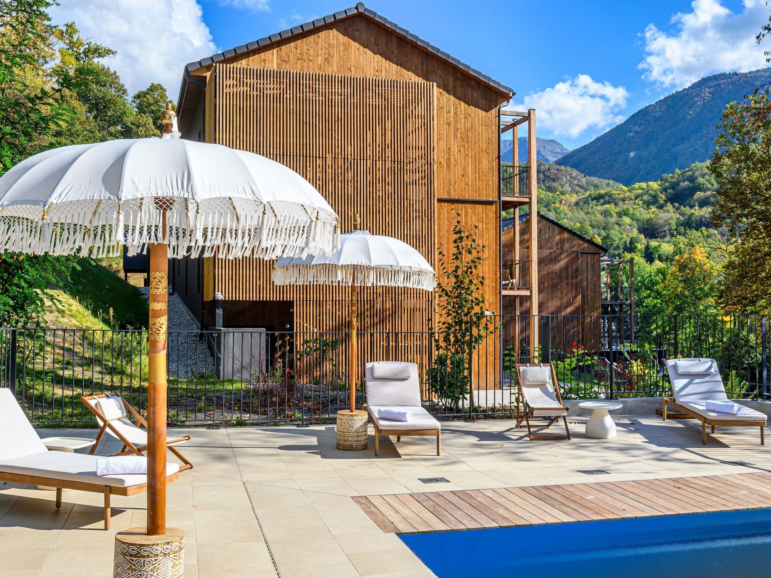 Un edificio de madera con piscina, sillas de descanso y sombrillas.