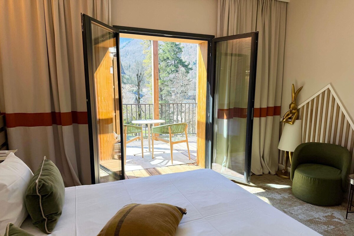 A hotel room with a bed, a green armchair, a lamp, and a view of a balcony with a table and chairs.