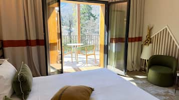 Ein Hotelzimmer mit Bett, einem grünen Sessel, einer Lampe und Blick auf einen Balkon mit Tisch und Stühlen.