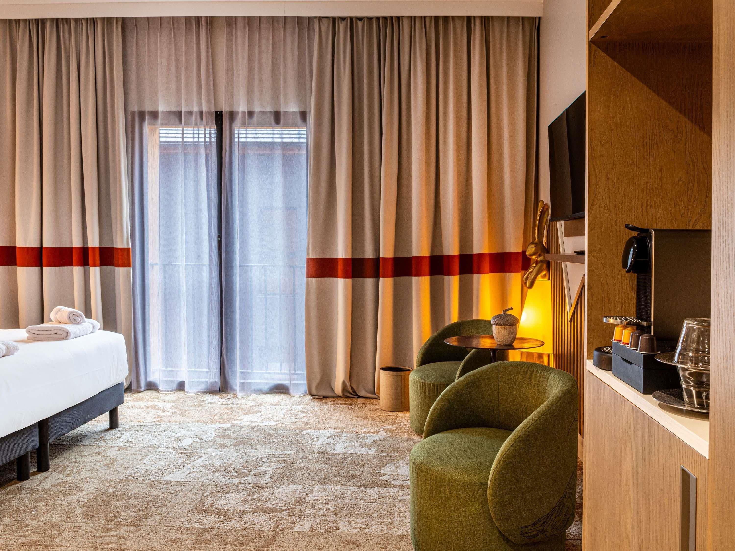 A hotel room with a bed, a green armchair, a coffee machine, and a television on a wooden cabinet.