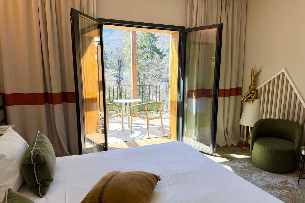 Ein Hotelzimmer mit Bett, einem grünen Sessel, einer Lampe und Blick auf einen Balkon mit Tisch und Stühlen.