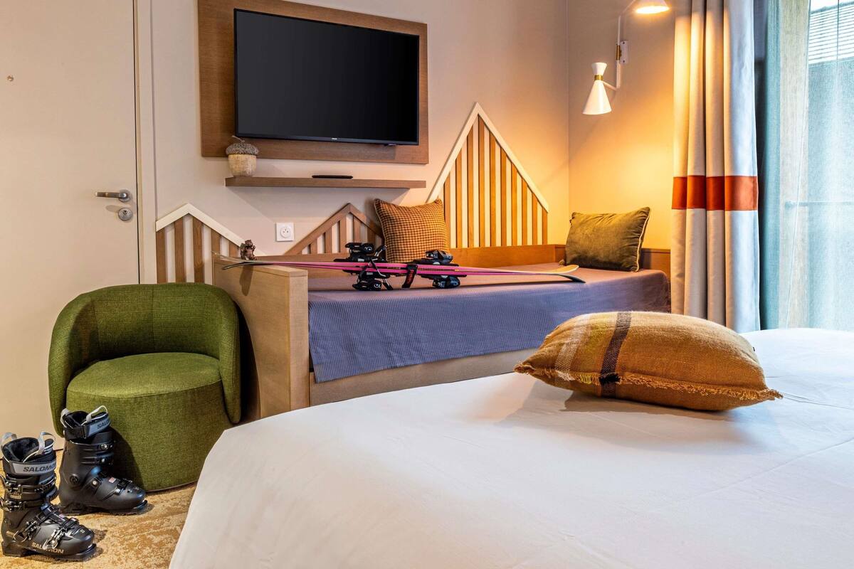 Ein Hotelzimmer mit Bett, Wandfernseher, Sessel und einem Paar Ski.