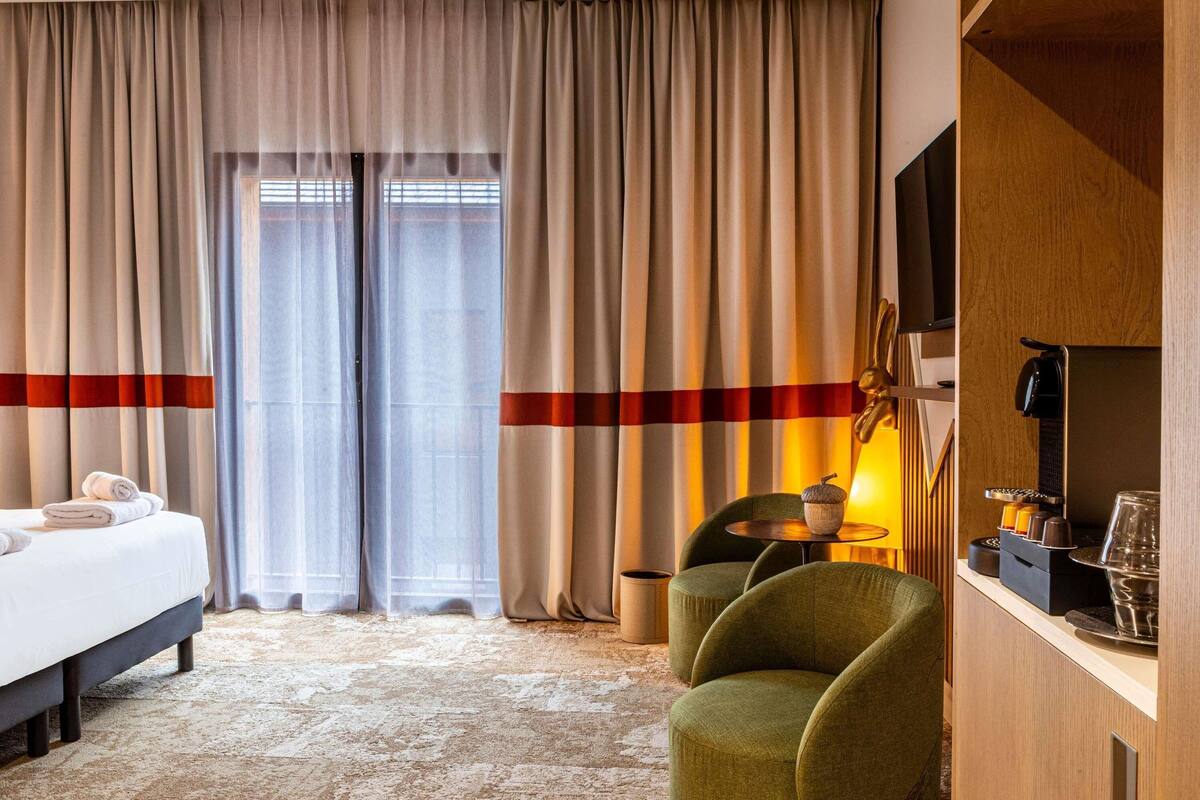 Ein Hotelzimmer mit einem Bett, einem grünen Sessel, einer Kaffeemaschine und einem Fernseher auf einer Holzkommode.