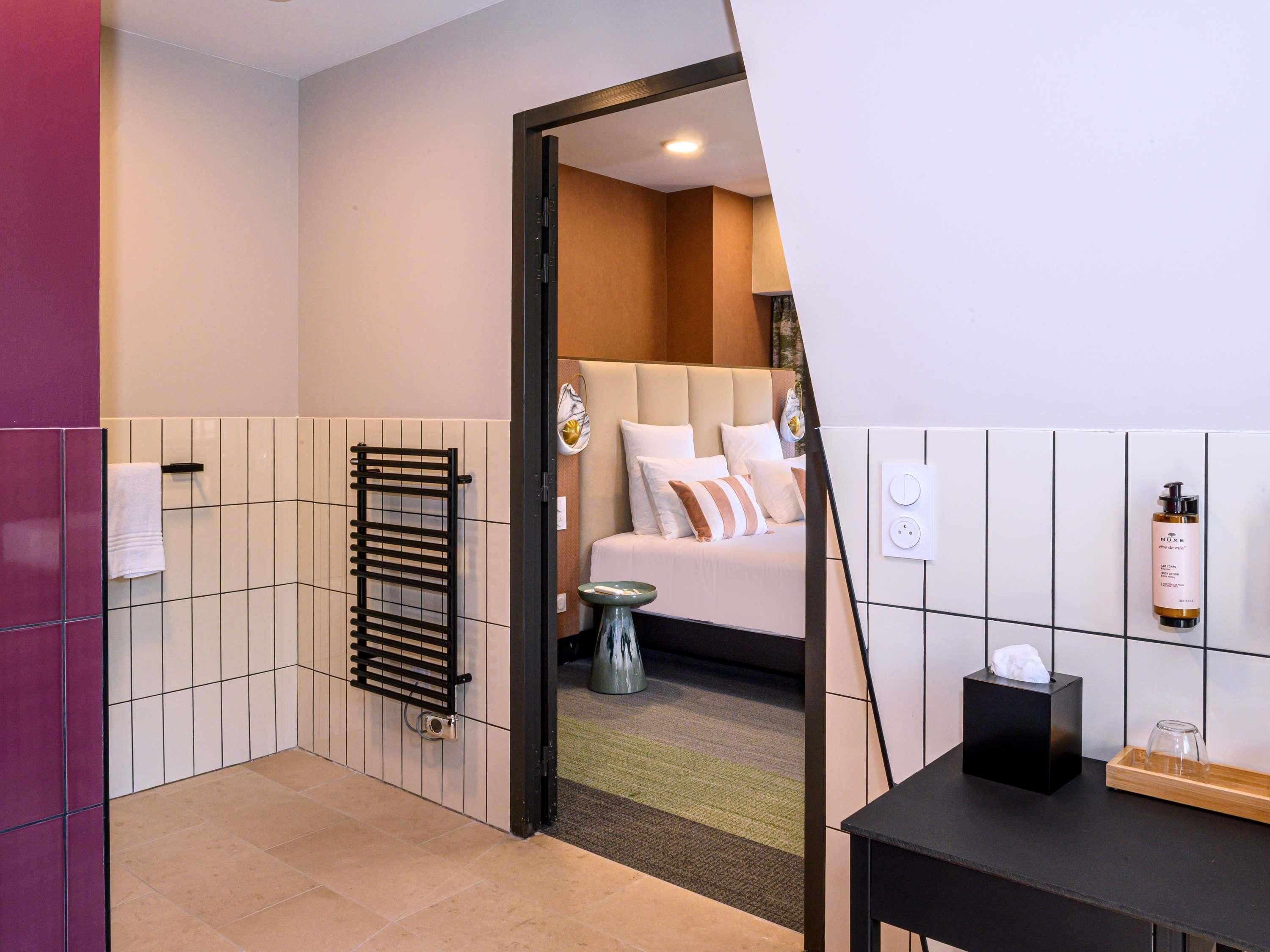 Ein Hotelzimmer mit Bett, Handtuchhalter, kleinem Tisch, einer Flasche Nuxe Hautpflegeprodukt und einer Steckdose.